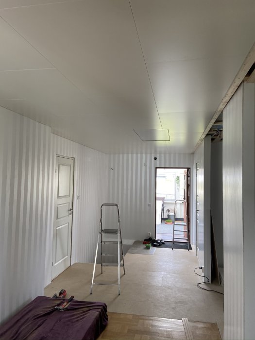 Renoveringsrum med omonterat innertak och verktyg, stege i förgrunden och öppen dörr till trädgården.