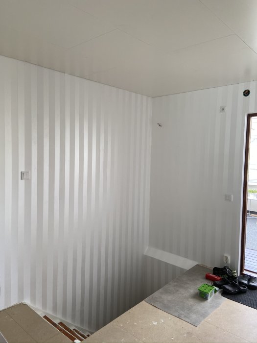 Ett rum under renovering med omonterat innertak och väggar tapetserade i randigt mönster, verktyg på golvet.