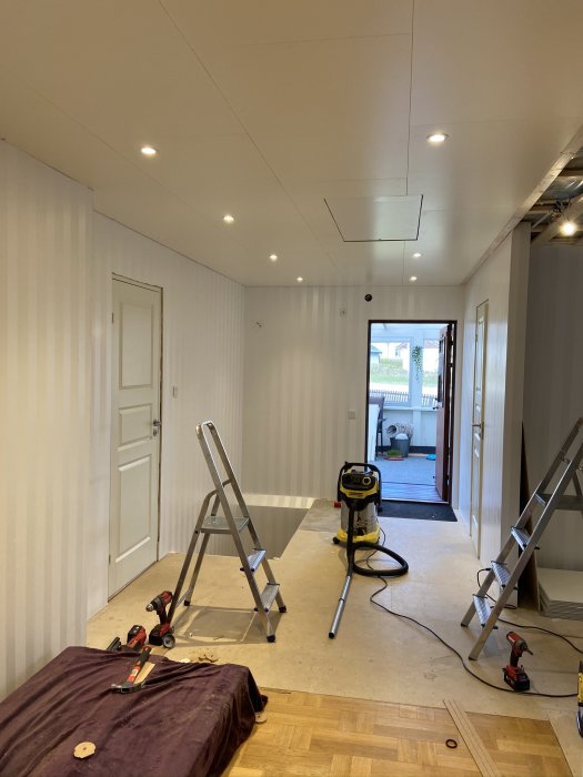 Renoverad inomhusmiljö med nytt innertak, utskurna hål för spotlights, stege, verktyg och dammsugare på golvet.