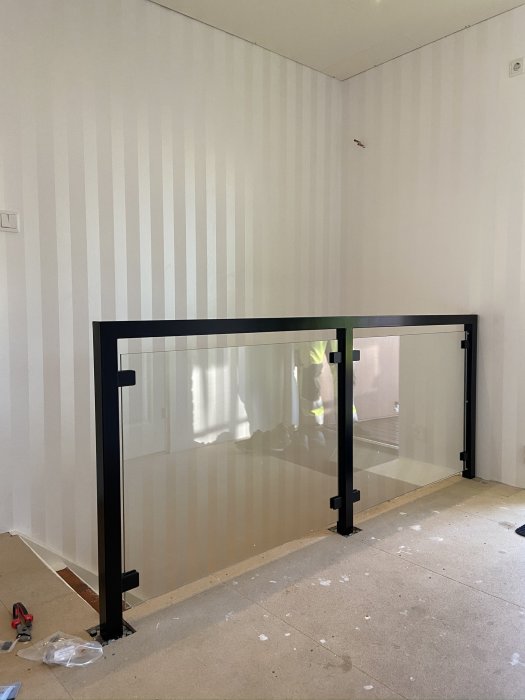 Svart räcke med glasskiva som väntar på installation i ett rum med vit randig tapet.