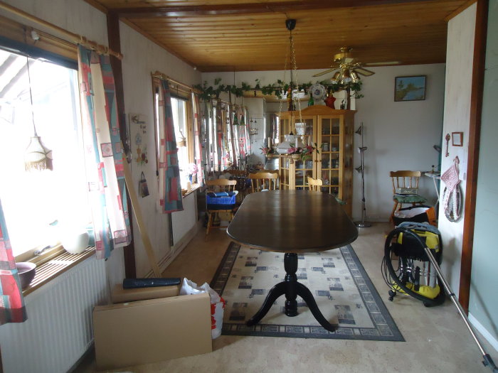 Ljust kök med ett ovalt bord i förgrunden, vitrinskåp och stolar i bakgrunden, fönster med gardiner och dekorationer.