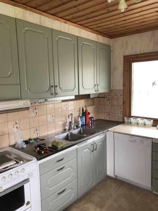 Kök före renovering med gröna skåp, kakel och verktyg på diskbänken.