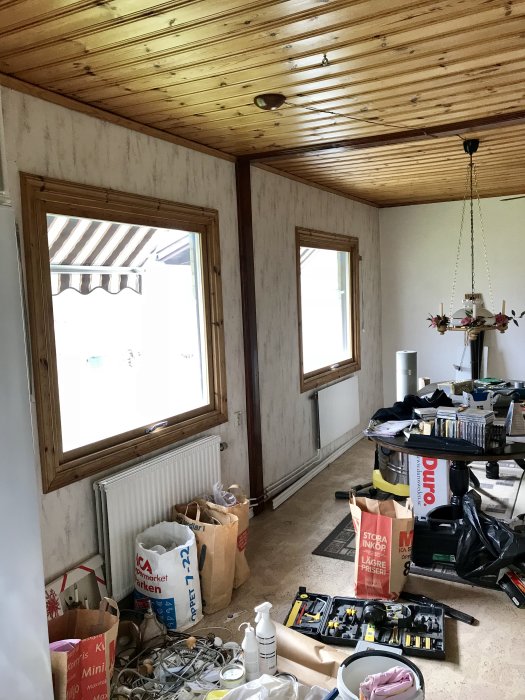 Ett rörigt rum redo för renovering, med utspridda verktyg och byggmaterial, träpaneltak och tapetserade väggar.