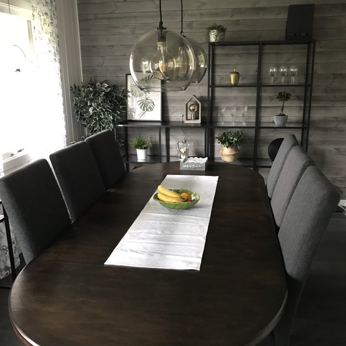 Nyrenoverat köksmatsal med mörkt träbord, grå stolar och modernt hyllsystem mot trävägg.