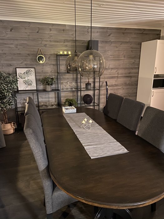 Ett nyligen färdigställt matrum med träbord, gråa stolar och designbelysning mot en vägg med träpaneler.