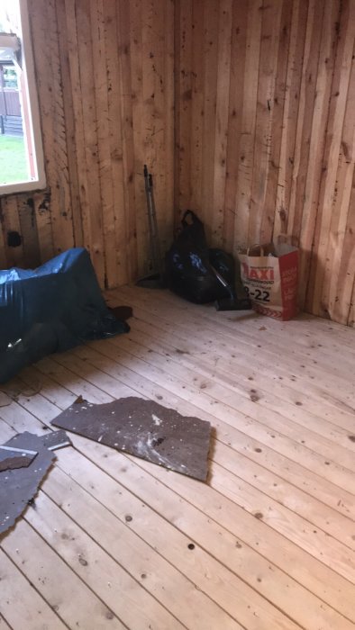 Ett hörn av ett tomt rum med träväggar och golv, rivningsavfall och en säck på golvet.