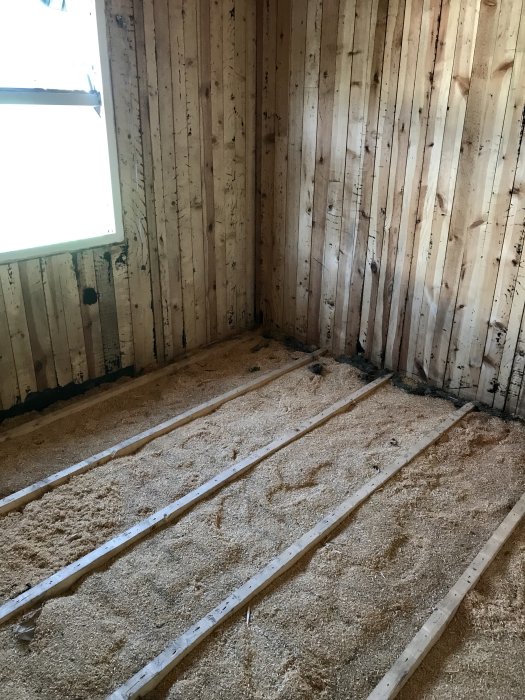 Tomt rum med sågspån på golvet, träväggar och tomma golvbjälkar synliga efter renovering.