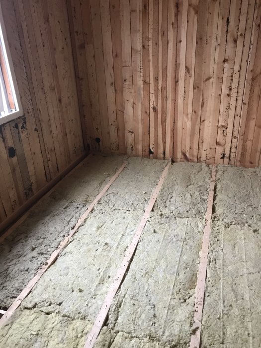 Isolering av mineralull utlagd mellan träreglar på ett golv i ett rum med träväggar och fönster, redo för golvspånskivor.