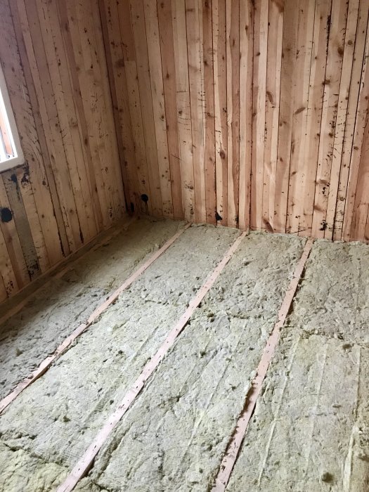 Isolering av mineralull mellan träreglar på ett golv i rum med träväggar, förberett för golvspånskivor.