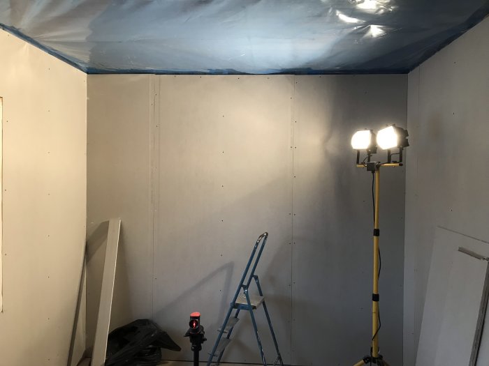 Inredning under renovering med oskadade gipsskivor på vägg och byggplast på taket, arbetslampor och stege.