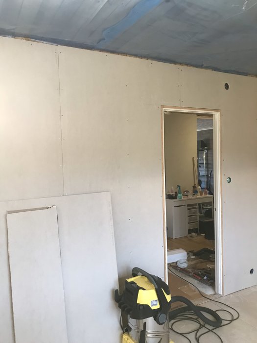 Renoveringsarbete med väggar klädda i gips och synlig byggplast i taket, dammsugare i förgrunden.