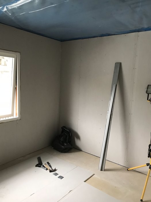 Ett rum under renovering med oslipade gipsväggar och blå byggplast på taket.