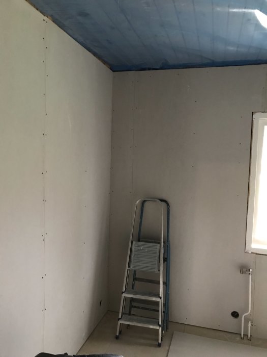 Vägg i rum med monterade gipsskivor och tak täckt av byggplast, stege står framför.
