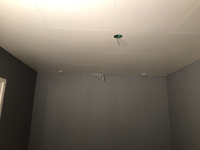 Ett nyttakserat rum med gråa tapetserade väggar och vitt innertak med synliga skarvar samt oinstallerade taklampor.