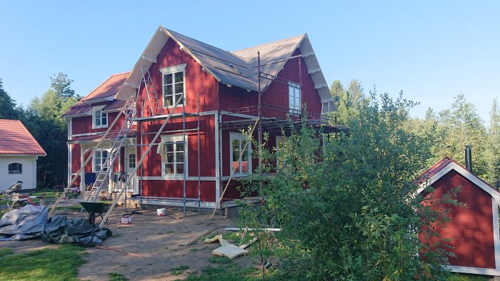 Byggplats med rött hus och pågående panelspikning, byggnadsställning och byggmaterial på marken.