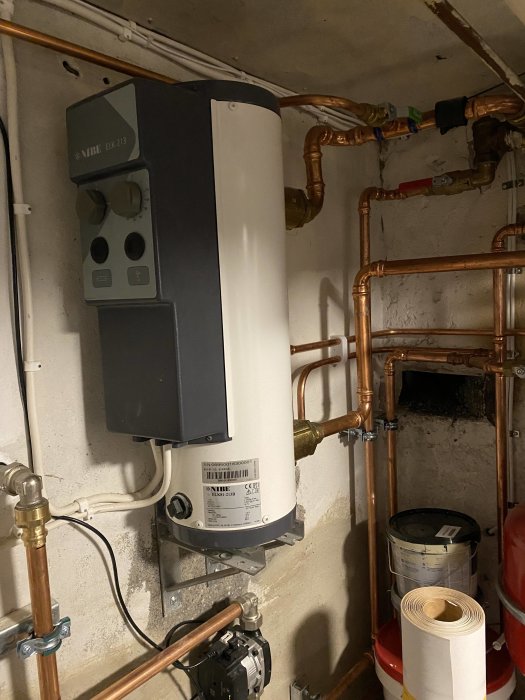 Värmepump och kopparledningar för golvvärme i ett fördelarskåp, med röd kran och cirkulationspump.