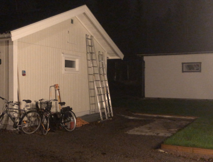 Husets kortsida med en stege lutad mot väggen, grus och cyklar framför, öppen yta för planerad carport.