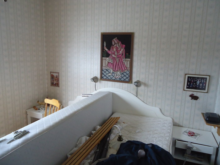 Sovrum med gammaldags tapet, omonterad säng, litet bord, och väggdekorationer.