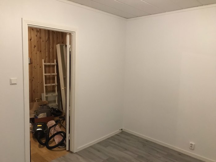 Renoverat rum med nytt laminatgolv och vita väggar, ny innerdörr på vänster och renoveringsmaterial.