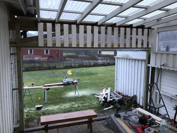 Pågående byggnation av vägg med horisontell träpanel under ett genomskinligt tak, verktyg och material synliga.