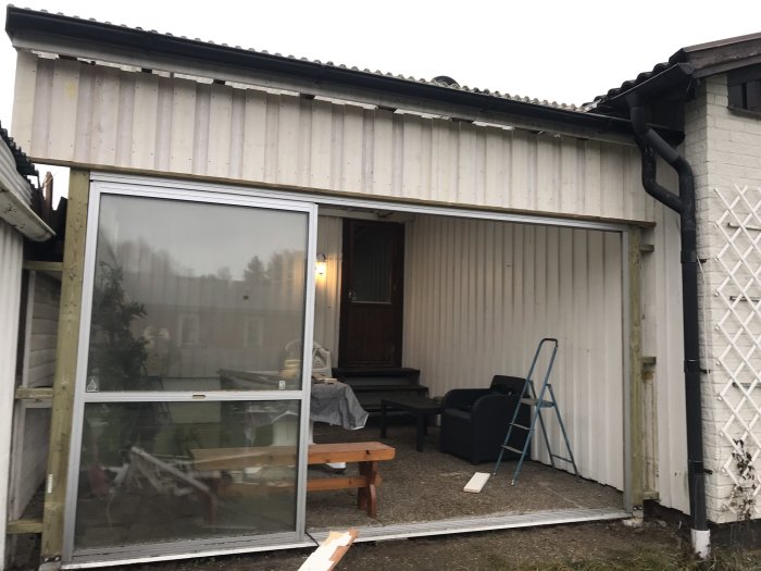 Husfasad med öppen garageport och utan panelbeklädnad, stegar och byggmaterial synliga.
