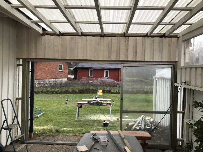 Vit målad uteplats under konstruktion med genomskinligt tak och trädgårdsutsikt.
