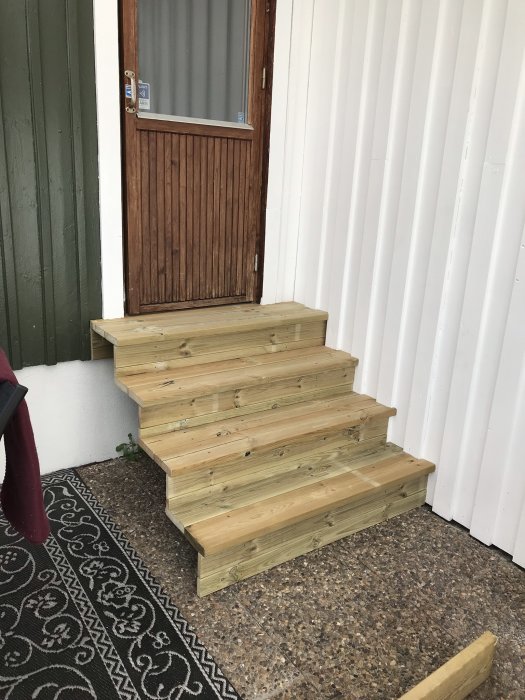 Nybyggda trätrappsteg framför en brun dörr till ett uterum mot vit vägg.