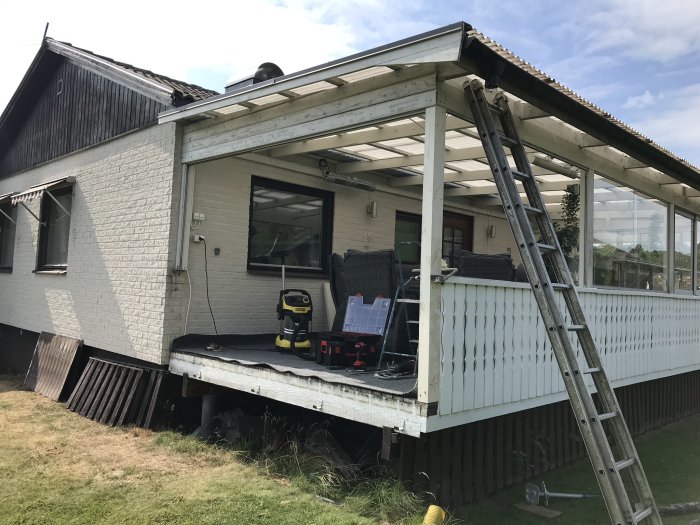 Ställning använd som stöd för ett delvis demonterat träveranda under renovering vid ett vitmålat hus.