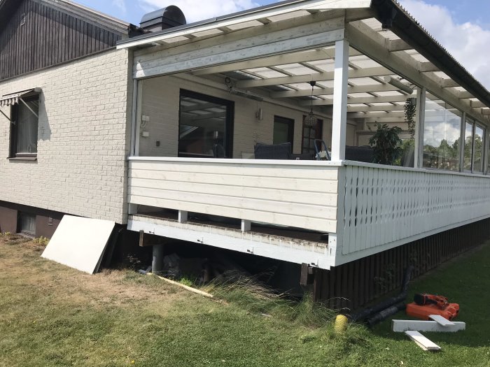Vit veranda med nytt räcke och panel på ett hus, delvis renoverat, med verktyg på gräset.