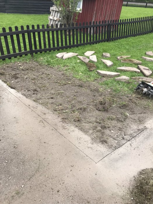 Nygrävd jord där en gammal rabatt varit, med stenplattor utspridda på gräset vid ett staket.