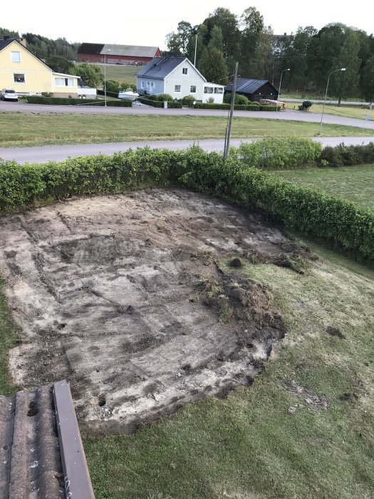 Schaktad trädgårdsyta med synliga ojämnheter och jord, framför en häck med hus och lantlig miljö i bakgrunden.