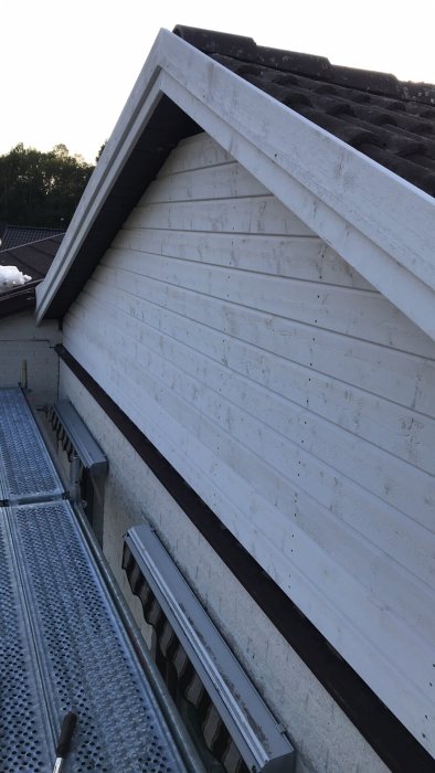 Ny liggande vit träpanel på gaveln av ett hus, med tak och stege i förgrunden.