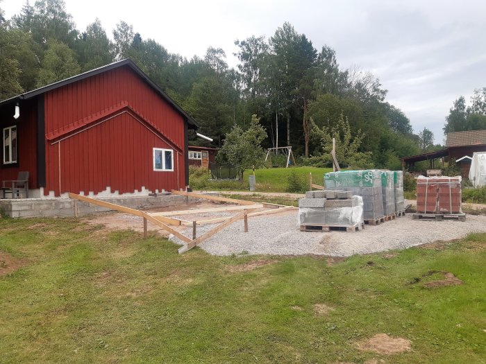 Röd lada under renovering med materialpaket för byggprojekt på grusgrund framför byggnaden.