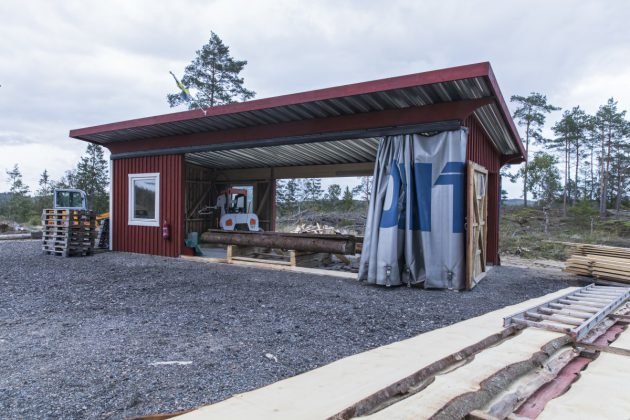 Enklare såghus med röda väggar, stor öppning och sadeltak, omgivet av grusmark och skogsmiljö.