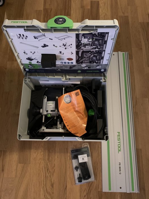 Festool OF1010 överfräs i öppen låda med tillbehör och justeringsverktyg, samt ett Festool-skenpaket på golvet.