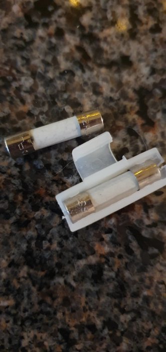 Två säkringar ligger på en marmorlik yta, en hel och en med synligt bränt element.