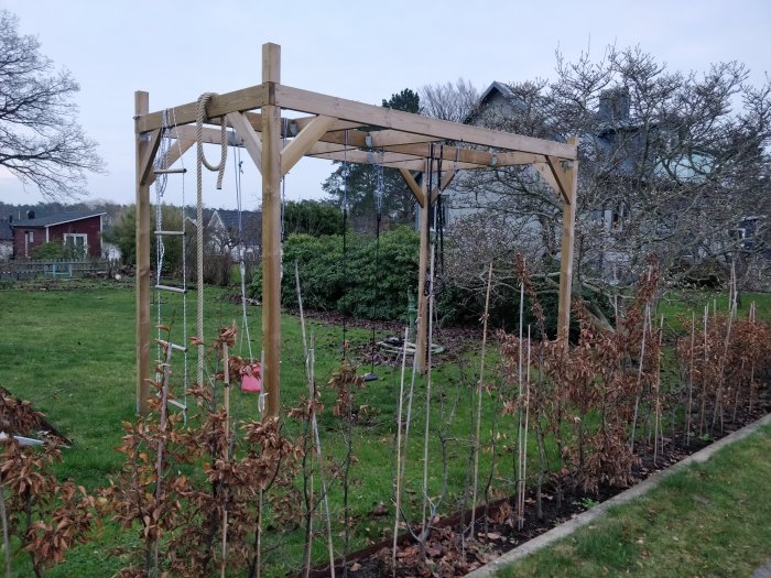 En oavslutad träkonstruktion av en klätterställning med saknad monkeybar-del och tvärstag, förankrad med betongblock.