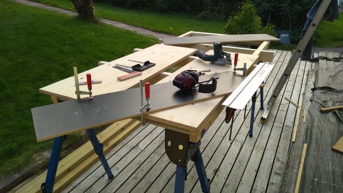 Två hopfällbara Kregbockar som stödjer en konstruktion av kapade plywooddelar och verktyg på ett utomhusbygge.
