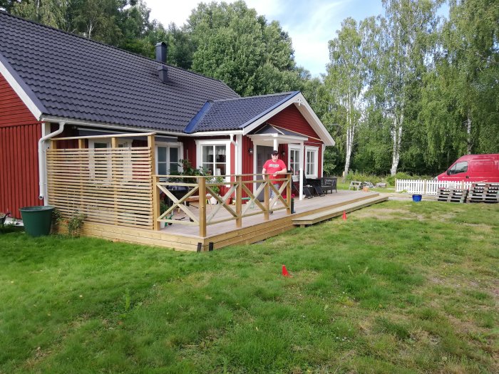 Person standing proudly on a newly built wooden deck of a renovated red house with a veranda and white picket fence.