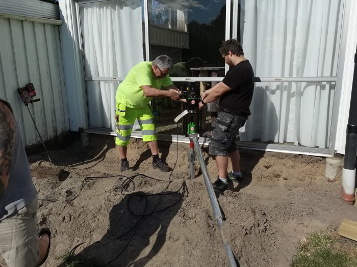 Två män använder en maskin för att installera en markskruv vid en byggnad.