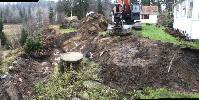 Grävmaskin vid en nyligen grävd grop nära ett hus, med högar av jord och grönska runtomkring.