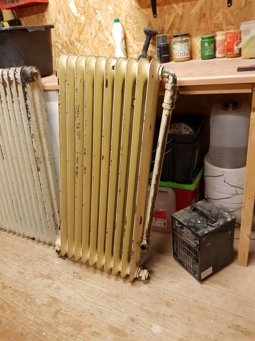 Begagnad golvstående radiator lutad mot en arbetsbänk i ett verktygsfyllt utrymme, indikerar renoveringsprojekt.