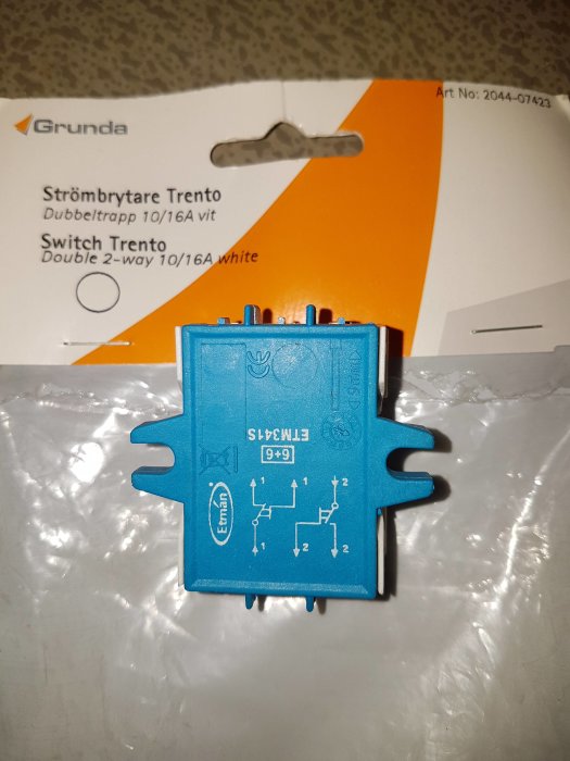 Blå strömbrytare med dubbeltrappfunktion och elschema, förpackning från Grunda i bakgrunden.