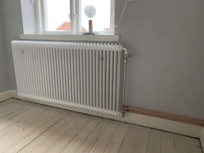 Vit radiator med nya termostatventiler i ett rum, trägolv och gråa väggar, under ett fönster.