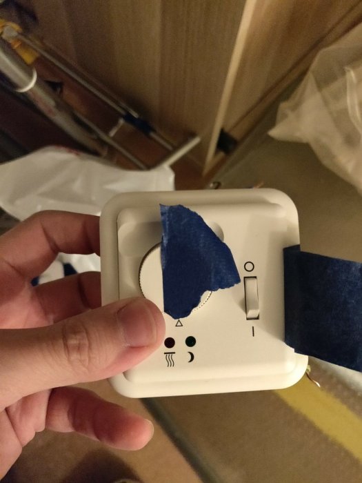 Gammal termostat täckt med blå tejp i en hand, mot bakgrund av ett rum under renovering.