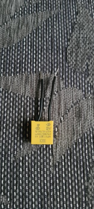 Kondensator med två svarta ledningar mot en texturerad tygbakgrund, använd vid installation av smart strömbrytare.