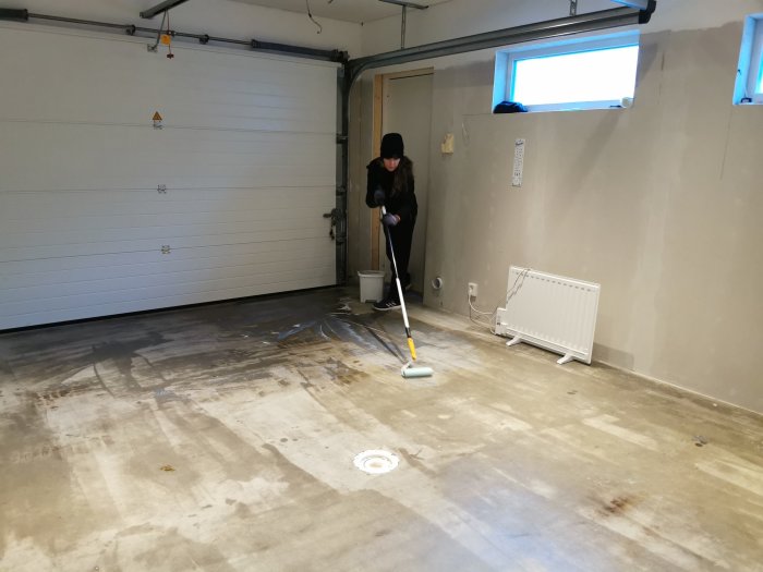 Person som tvättar ett betonggolv i ett halvfärdigt garage med vita väggar och garageport.