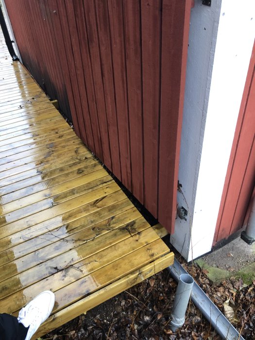 Öppen ränna med löv vid träaltan med lutning nära husvägg i dagsljus, begränsat utrymme för rörinstallation.