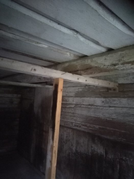 En timrad ladugårdsinteriör med kraftiga takbalkar och en vertikal stötta som möter en horisontell bjälke.