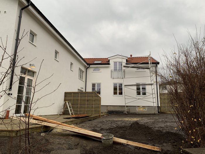 Äldre hus med modern tillbyggnad, synlig gräns mellan gammal och ny del, byggnadsställning och oanlagd trädgård.